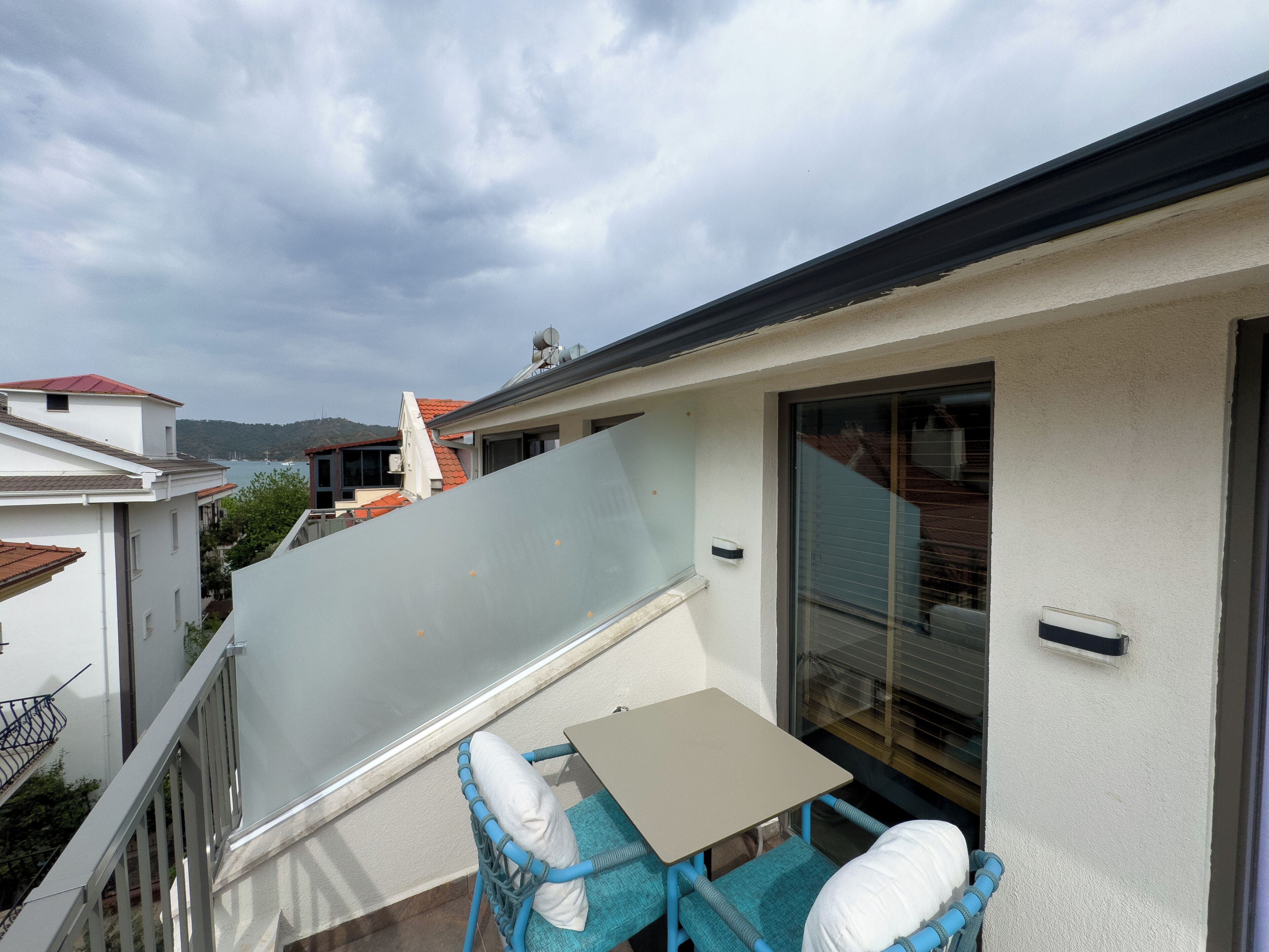 Ein Balkon mit Tisch und Stühlen, eine Glastür und Blick auf das Meer sowie Gebäude im Hintergrund.