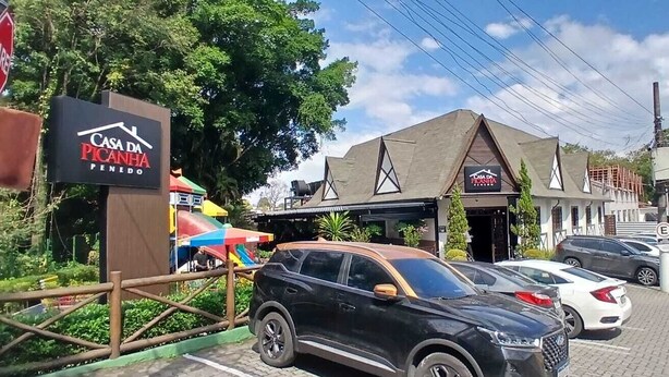 A street view with a sign for 'Casa da Picanha' and parked cars. - Alto da Colina Kitnetes - Penedo (RJ) (Penedo Itatiaia)