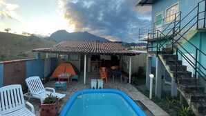 A swimming pool with lounge chairs, a tent, and a staircase leading to a building with a tiled roof. - Alto da Colina Kitnetes - Penedo (RJ) (Penedo Itatiaia)
