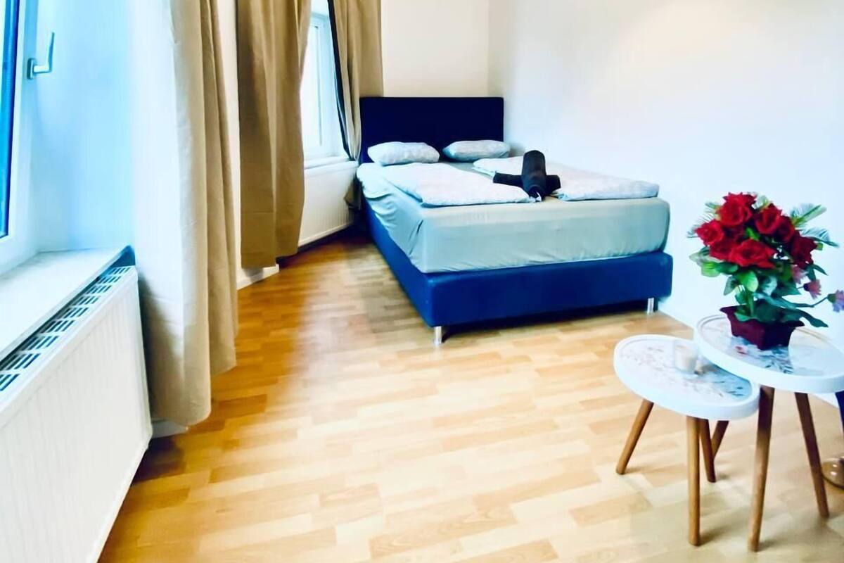 A bedroom with a bed, a radiator, a window with curtains, a wooden floor, and a small table with a flower vase.