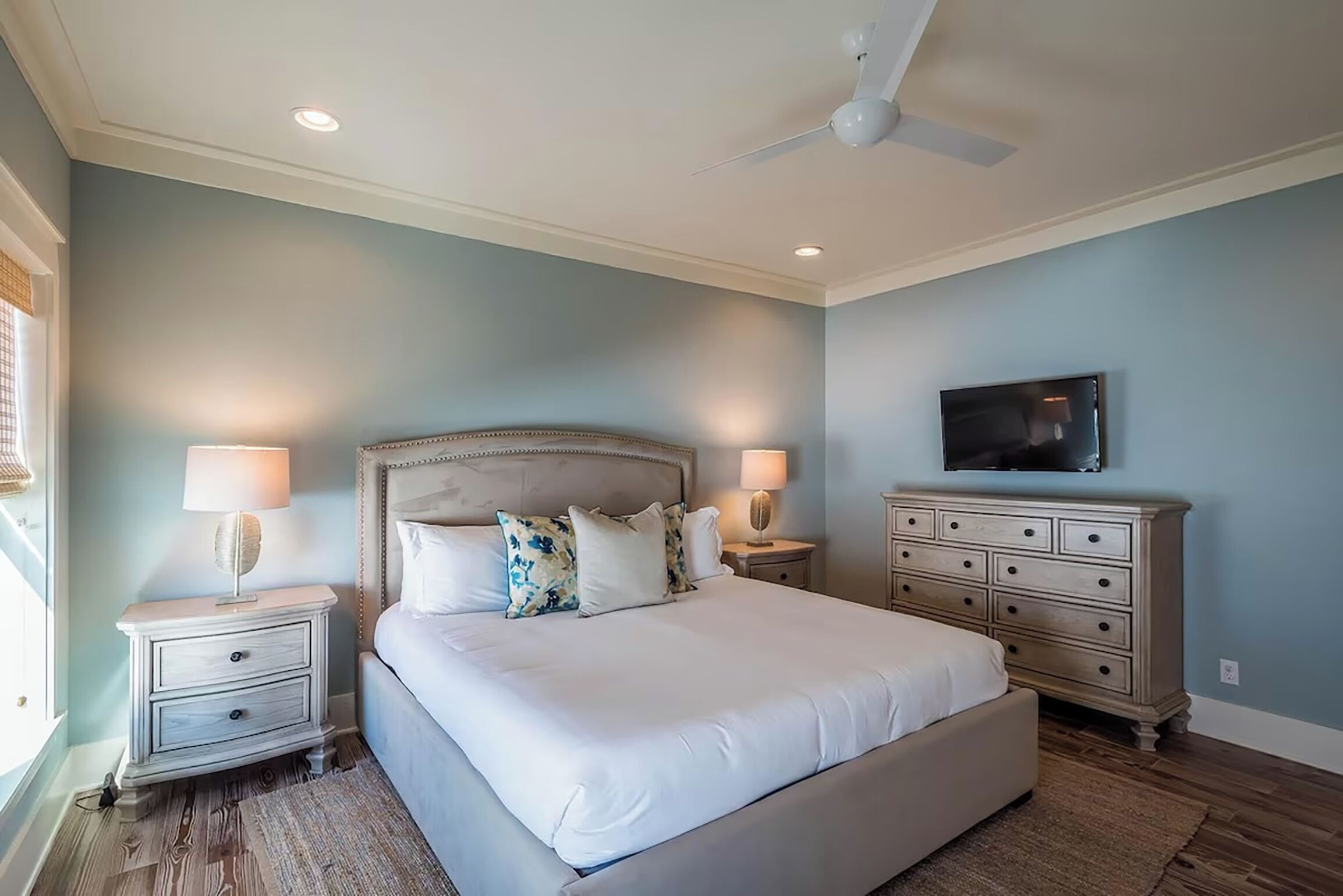 A bedroom with a large bed, two bedside tables, a ceiling fan, a TV, and a dresser.