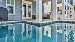 Une piscine avec un bord carrelé, un barbecue couvert et un patio abrité aménagé avec du mobilier d’extérieur.