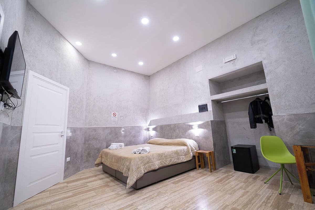 A modern bedroom with a bed, a small stool, a chair, a TV, and a coat rack.