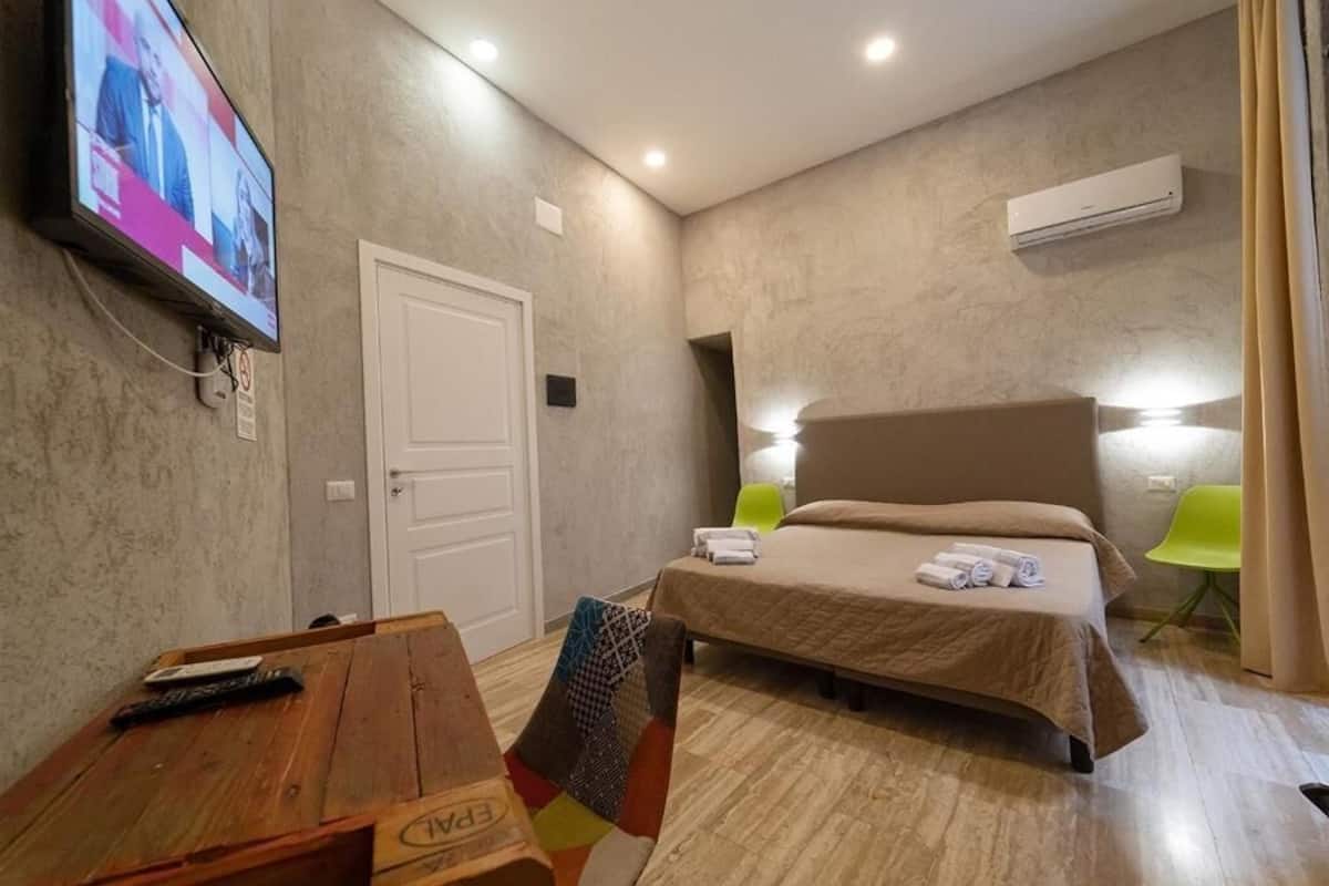 A hotel room with a bed, a desk with a chair, a TV mounted on the wall, and an air conditioning unit.