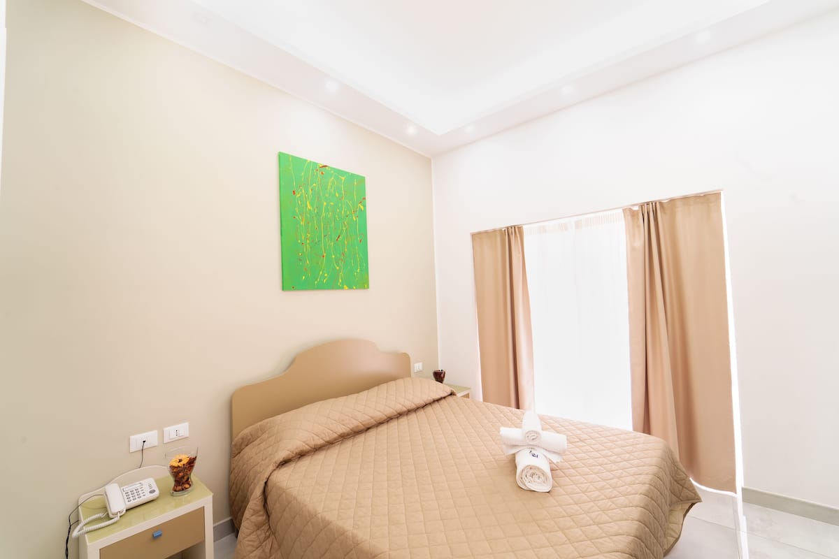 A hotel room with a bed, a nightstand with a phone, a glass of water, and a green abstract painting on the wall.