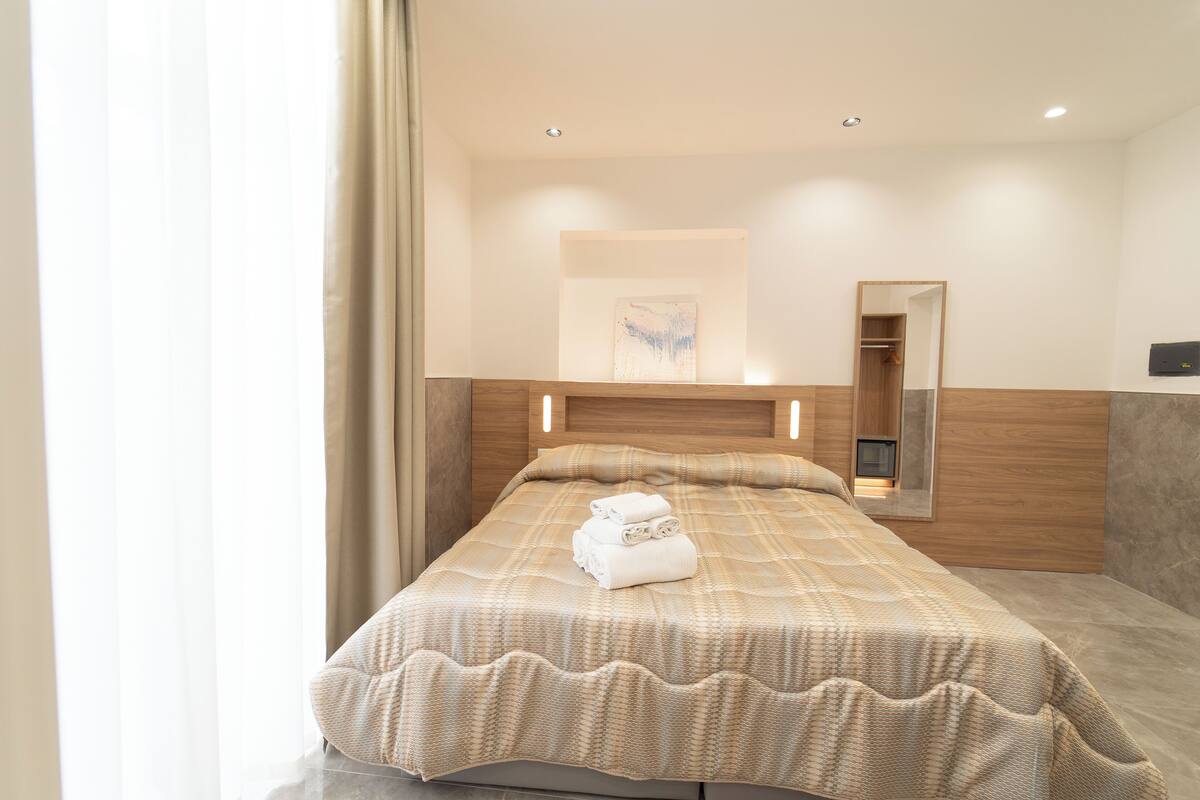 A neatly made bed with a checkered bedspread and rolled towels, a bedside table with a lamp, a mirror, and a wooden headboard.