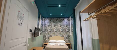 Une chambre d’hôtel étroite, comprenant un lit, une table de chevet, une petite table, une armoire et un miroir.