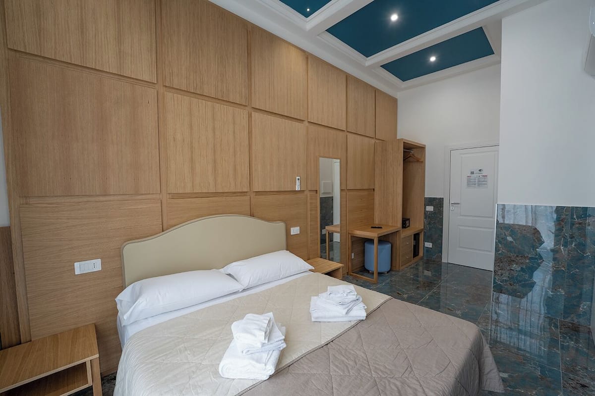 A hotel room with a large bed, a wooden wardrobe, a desk, and a bathroom with a marble floor.