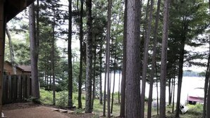 Una cabaña rústica con un sendero de tierra que lleva a una terraza de madera, rodeada de árboles altos y vistas a un lago.