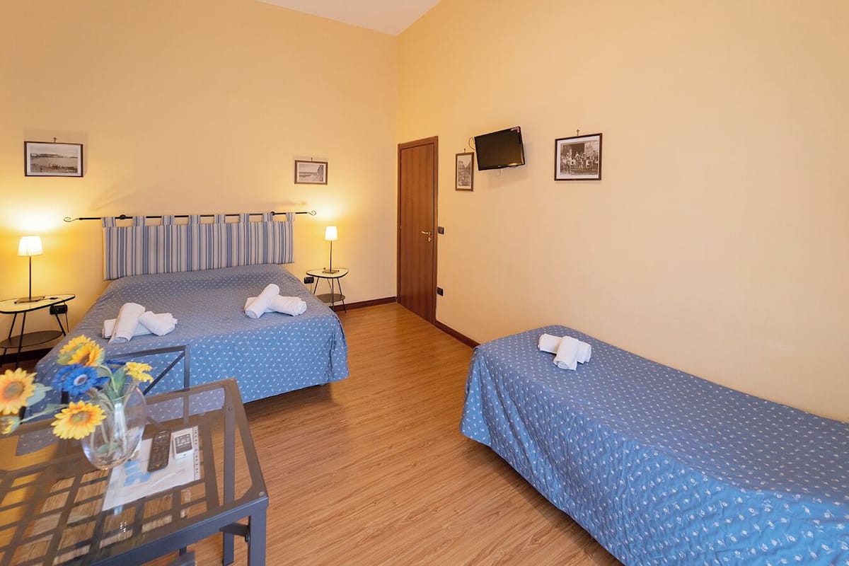 A hotel room with two beds, a small table with a vase of flowers, and a TV on the wall.