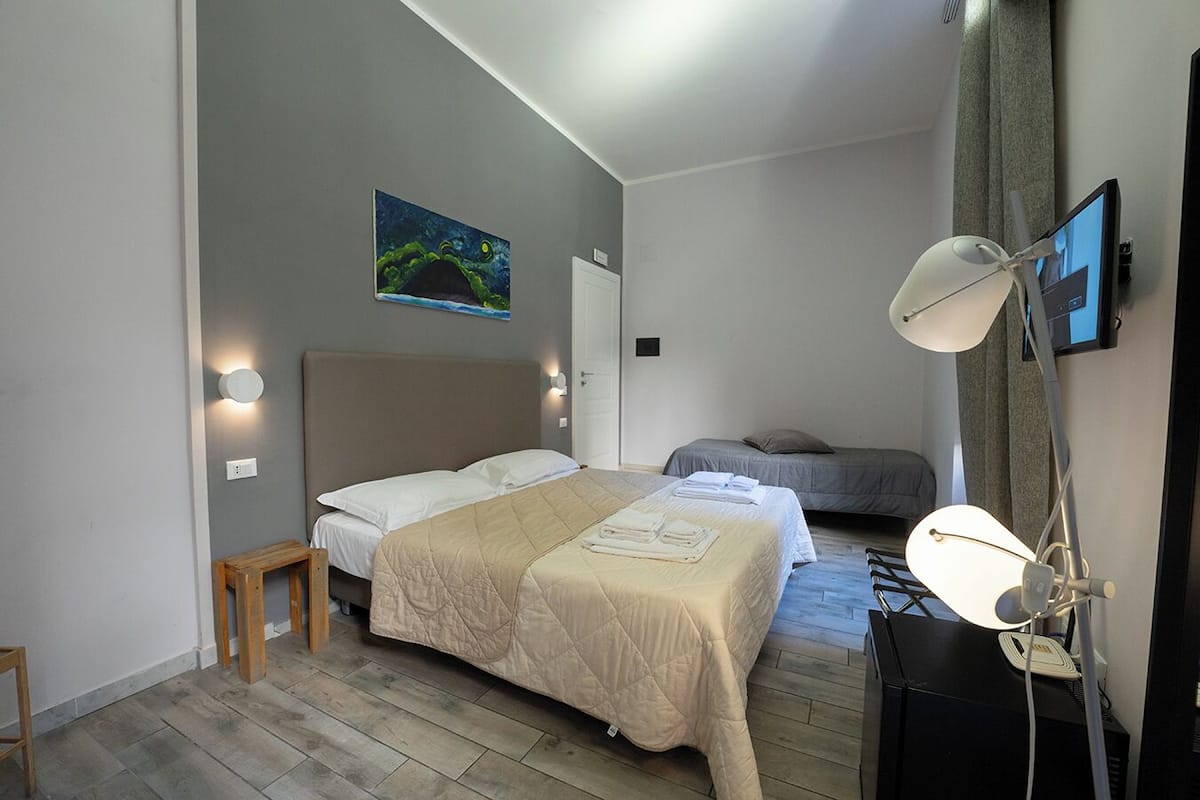 A modern bedroom with a large bed, a smaller bed, a desk with a lamp, and a painting on the wall.