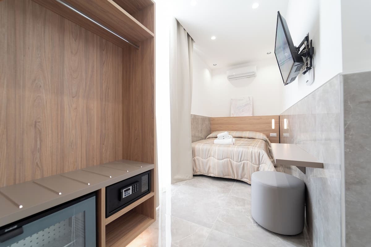 A modern hotel room with a wooden wardrobe, a bed with a beige bedspread, a grey ottoman, and a mounted flat-screen TV.