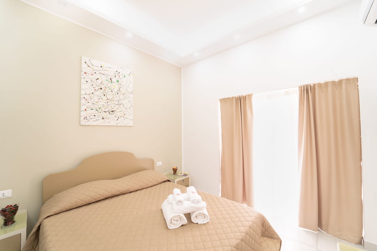 A hotel room with a bed, a nightstand, a painting, and a window with curtains.