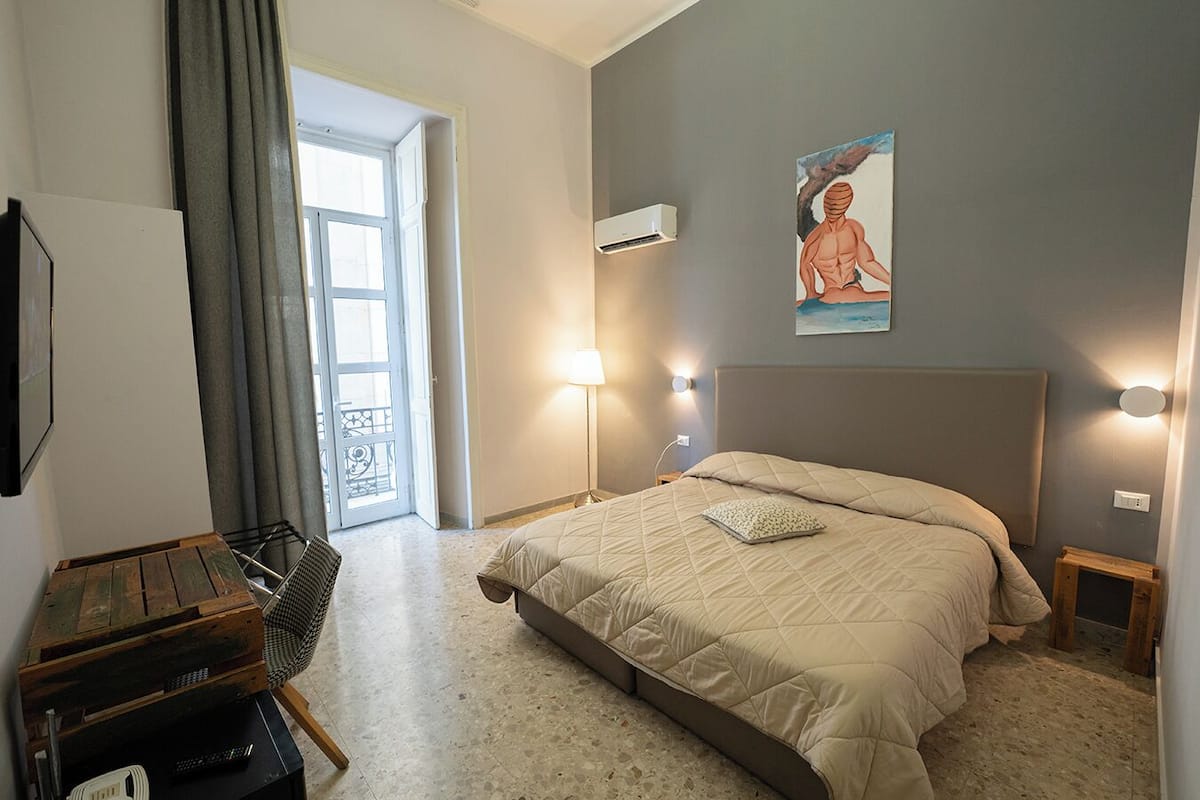 A bedroom with a bed, bedside lamps, a painting, and a view through an open door.