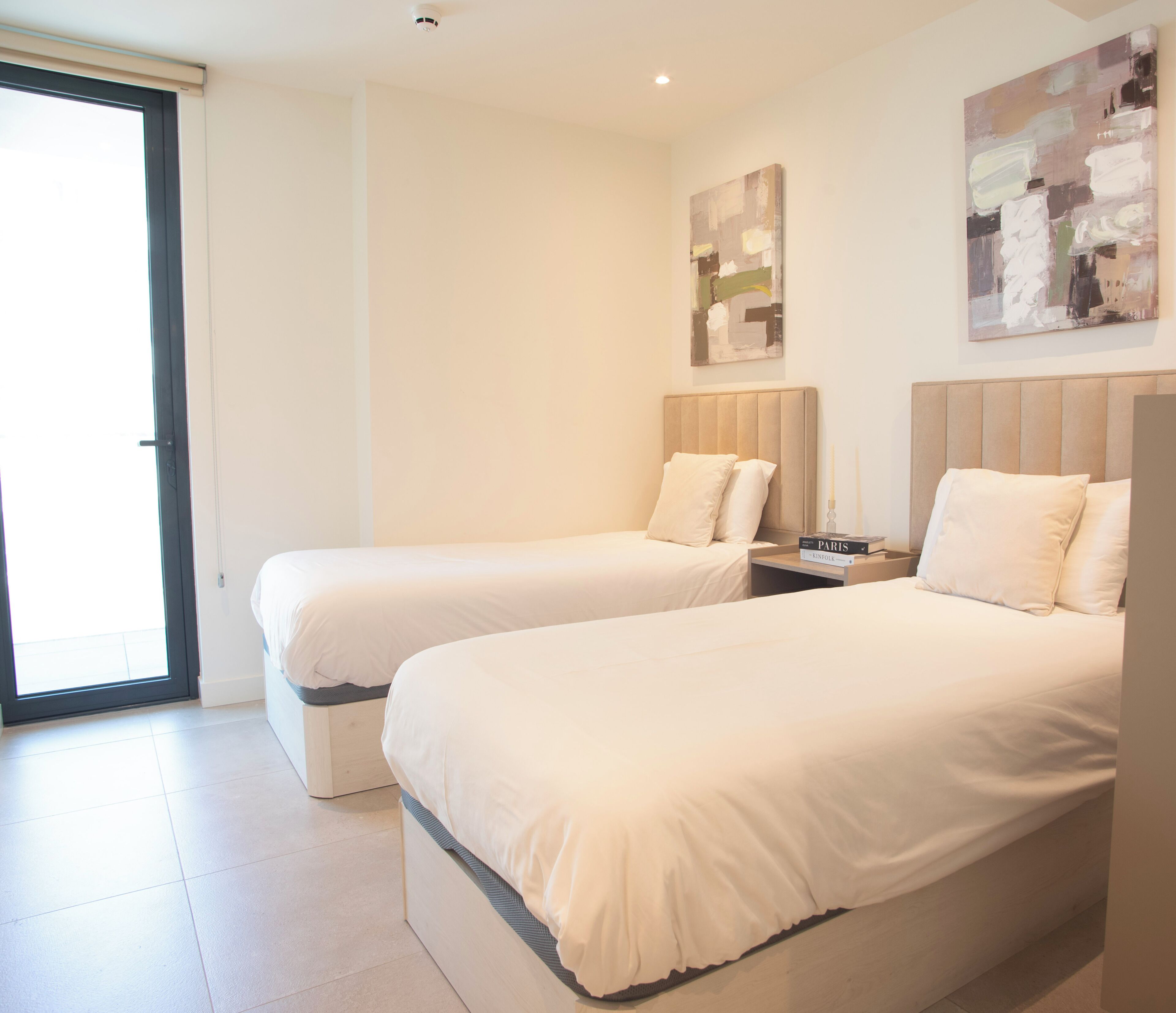 A hotel room with two beds, a nightstand, a lamp, and a sliding glass door.