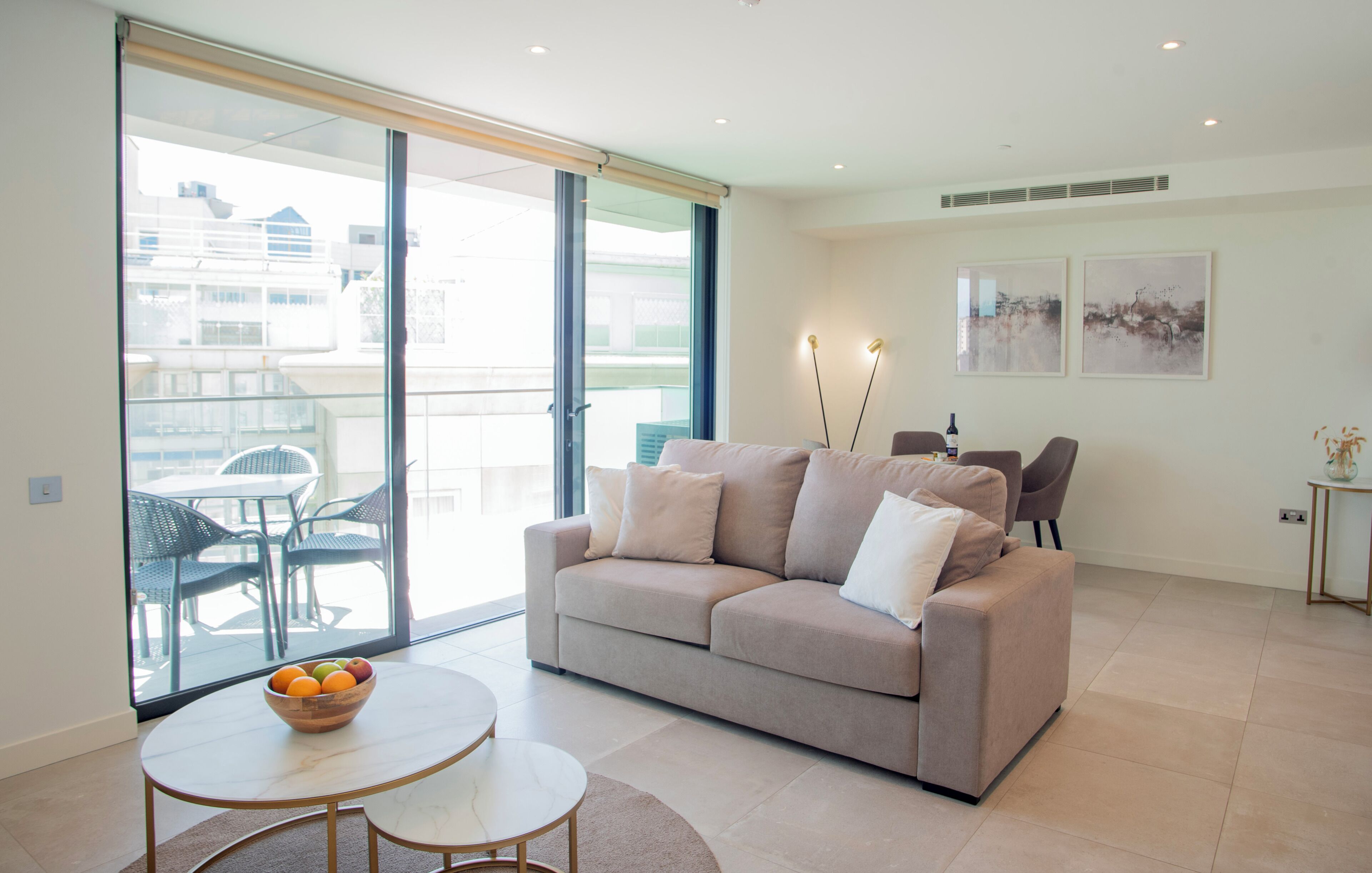 A modern living room with a sofa, a dining table with a fruit bowl, and a balcony with outdoor furniture.