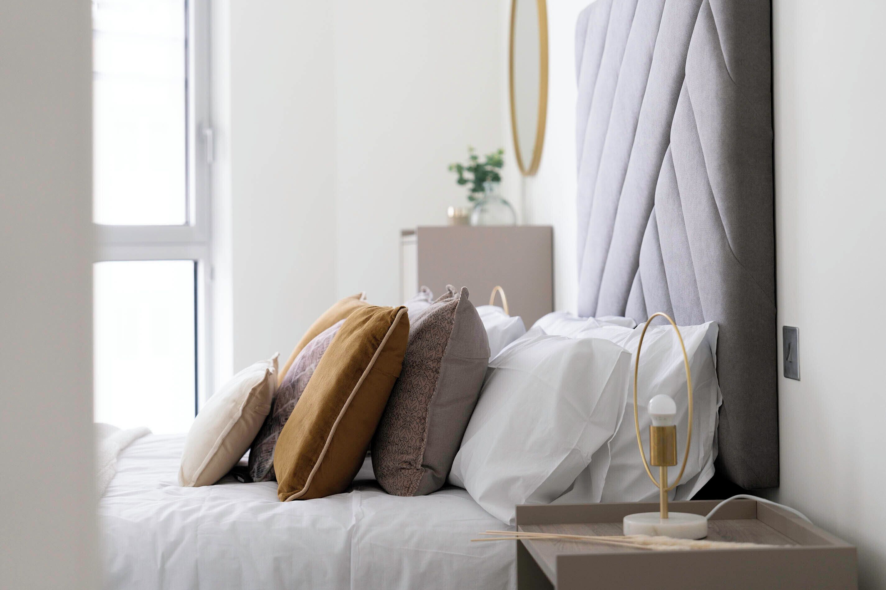 Un lit bien fait, avec du linge de lit blanc et plusieurs oreillers, une table de chevet avec une lampe et une fenêtre laissant entrer la lumière naturelle.