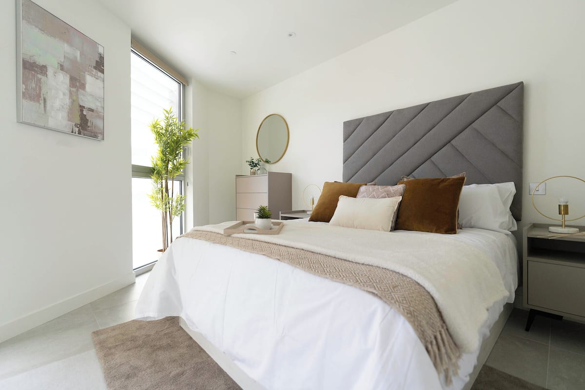 A modern bedroom with a large bed, a bedside table, a nightstand, a mirror, and a window with a view of greenery.