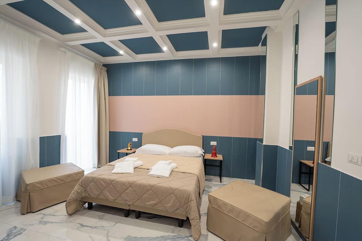 Une chambre d’hôtel avec un grand lit, deux tables de chevet, un miroir et une fenêtre avec des rideaux légers.
