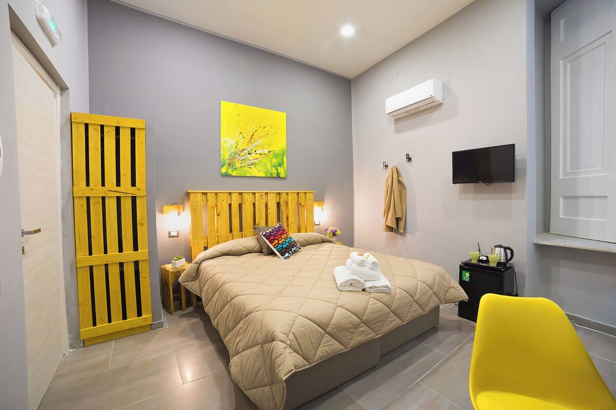 Une chambre à coucher avec un lit, une chaise jaune, un téléviseur et une petite table sur laquelle se trouve une bouilloire.