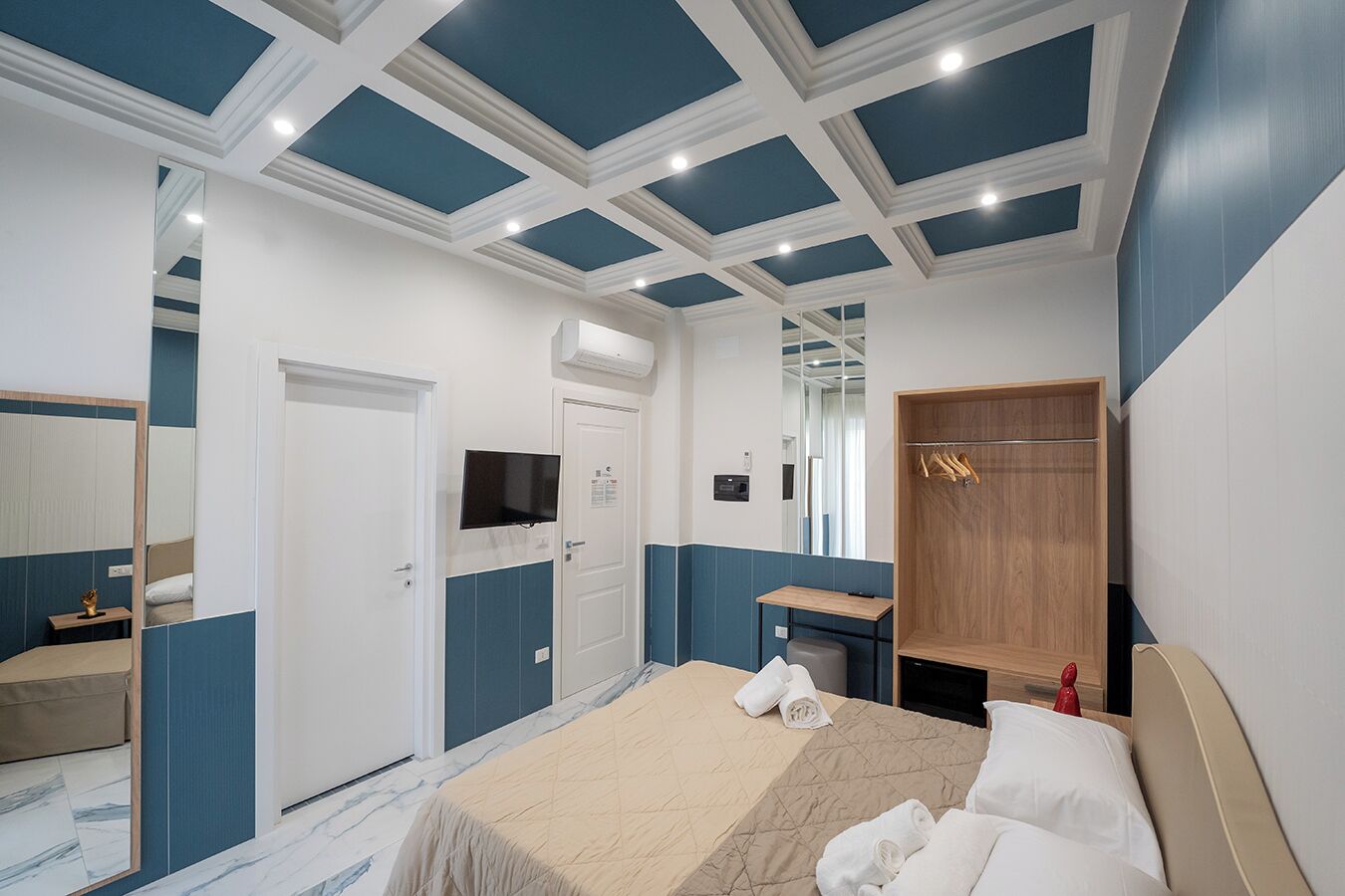 A modern hotel room with a bed, a wooden wardrobe, a desk with a chair, a television, and a mirror.