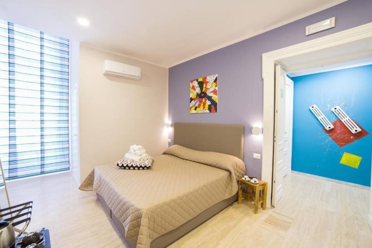 Une chambre à coucher comprenant un lit, une table de chevet, une fenêtre avec des stores et un mur orné d’un tableau abstrait aux couleurs vives.