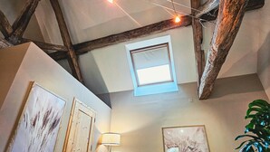 A rustic attic room with exposed wooden beams, a skylight, a black chair, a standing lamp, and framed artwork on the walls. - Family-Friendly 1BR w/ Garden View + Hot Tub (Étivey)