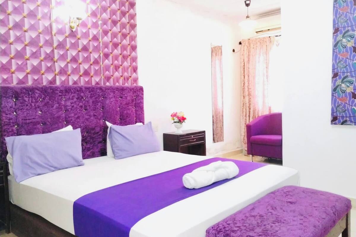Ein Schlafzimmer mit Bett, violettem Kopfteil, Nachttisch, Stuhl und Wand mit geometrischem Muster.