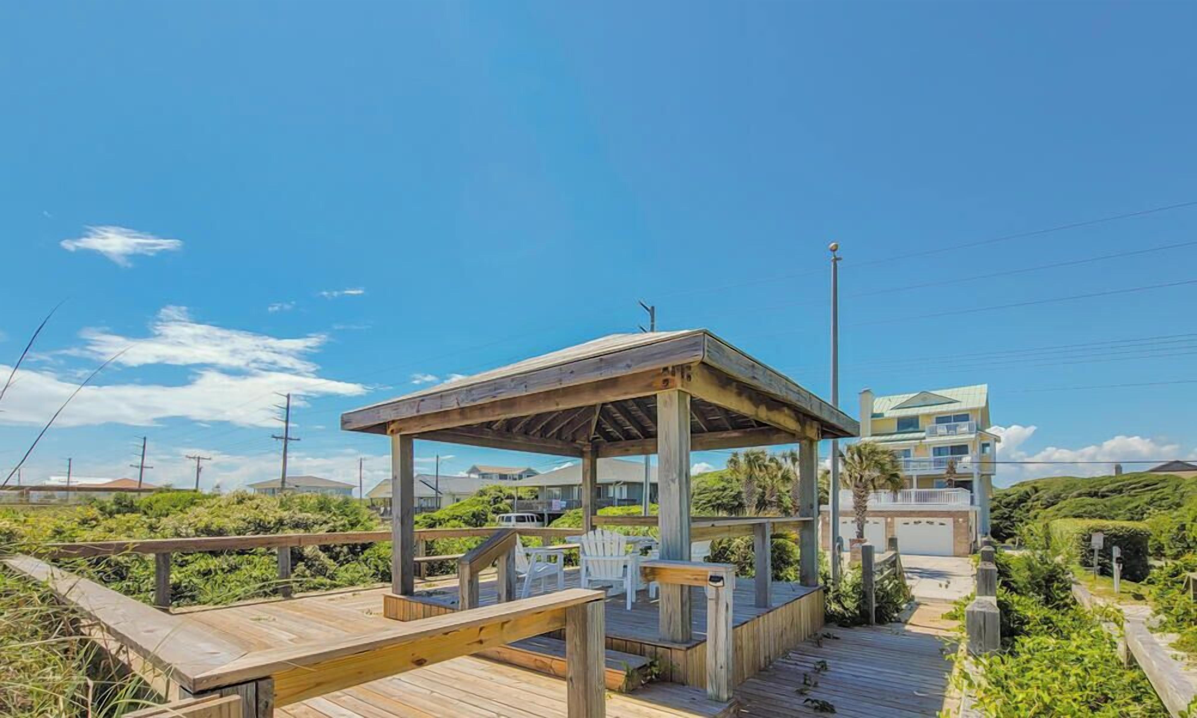 A wooden deck with a gazebo, white chairs, and a view of a coastal house.