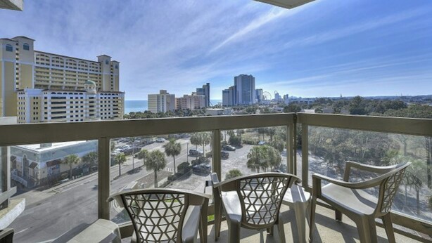 A balcony with two chairs, overlooking a cityscape with buildings and a clear sky. - Atlantis 704-2BR/2BA Myrtle Beach-Walk to Beach (Myrtle Beach)