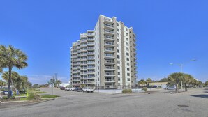 A tall, modern apartment building with multiple balconies, located at a street corner with palm trees and a clear blue sky. - Atlantis 704-2BR/2BA Myrtle Beach-Walk to Beach (Myrtle Beach)