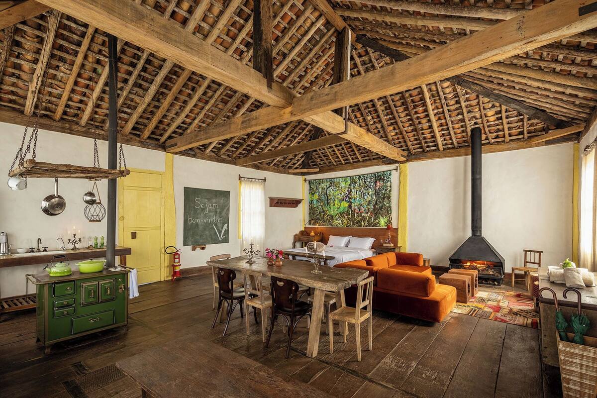 A rustic room with a high wooden ceiling, a fireplace, a dining area with wooden furniture, and a bed with an orange cushion.