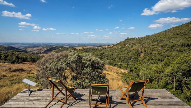 Eine Holzterrasse mit drei Holzstühlen, die auf eine reizvolle Hügellandschaft mit Bäumen und entfernten Hügeln unter blauem Himmel ausgerichtet ist.