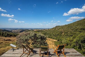 Une terrasse en bois avec trois chaises en bois, donnant sur un paysage pittoresque composé de collines, d’arbres et de montagnes lointaines, le tout sous un ciel bleu dégagé.