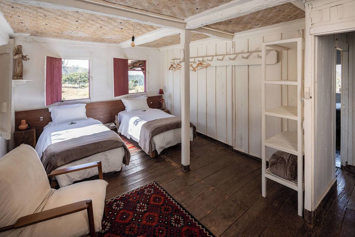 A room with two beds, a wooden floor, a red patterned rug, a white shelf, and a hanging light fixture.