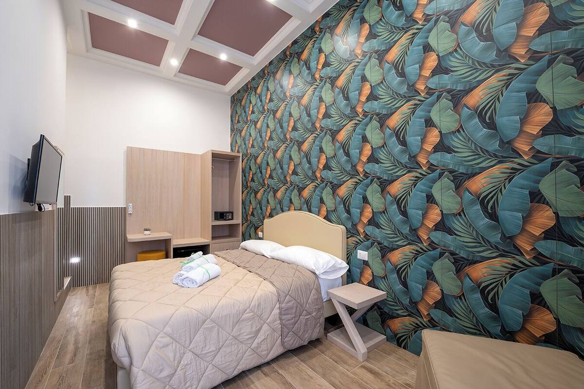 A hotel room with a bed, a TV, a wardrobe, a bedside table, and a wall with a leaf pattern.