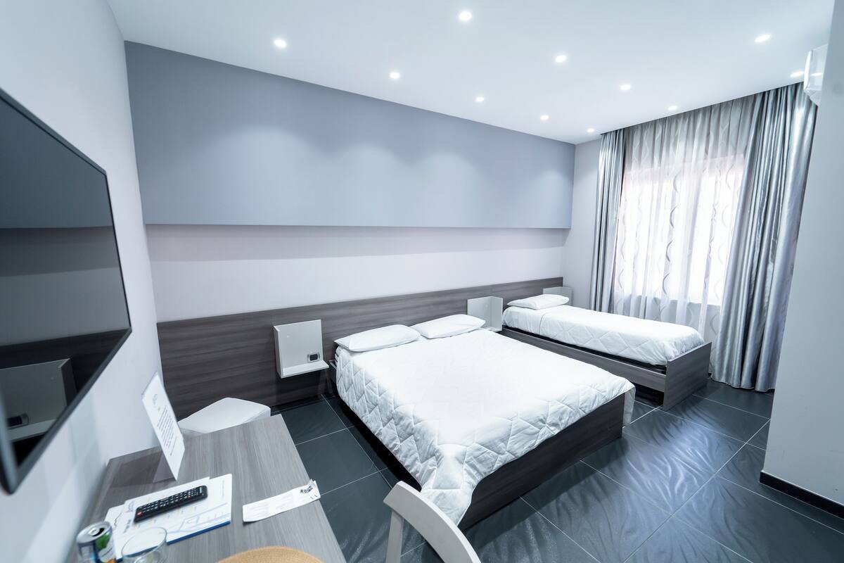A modern hotel room with a large bed, a smaller bed, a desk with a chair, a TV, and a window with curtains.