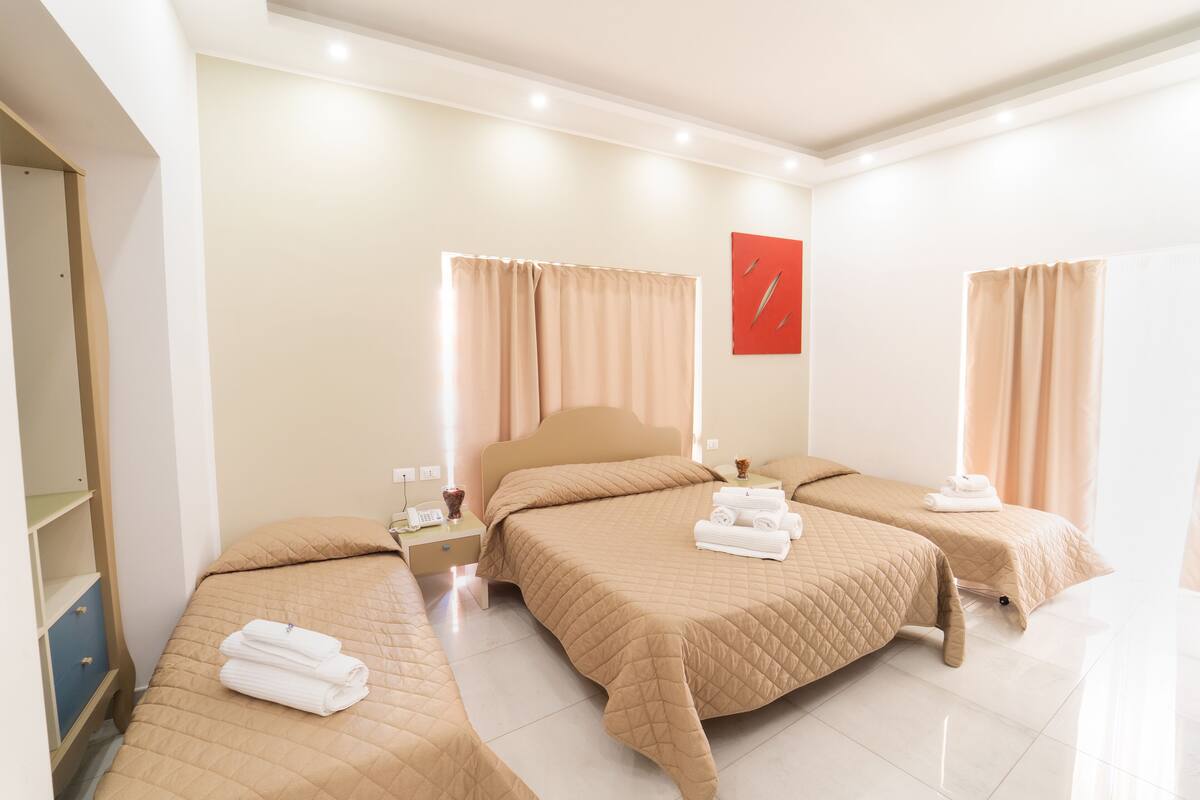 A hotel room with three beds, each with a beige quilted bedspread and neatly folded towels. There is a wooden wardrobe on the left, a nightstand with a lamp, and a red abstract art piece on the wall.