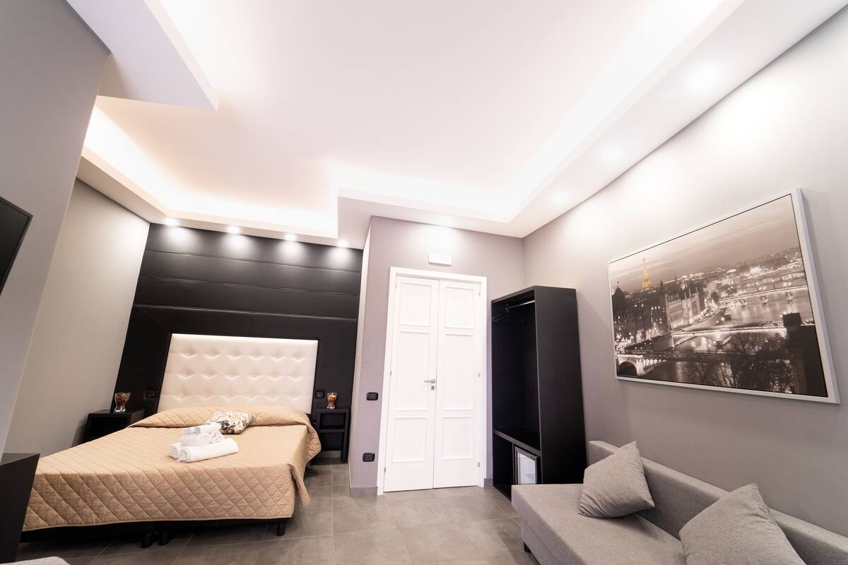 A modern bedroom with a large bed, a sofa, a TV, and a cityscape painting.
