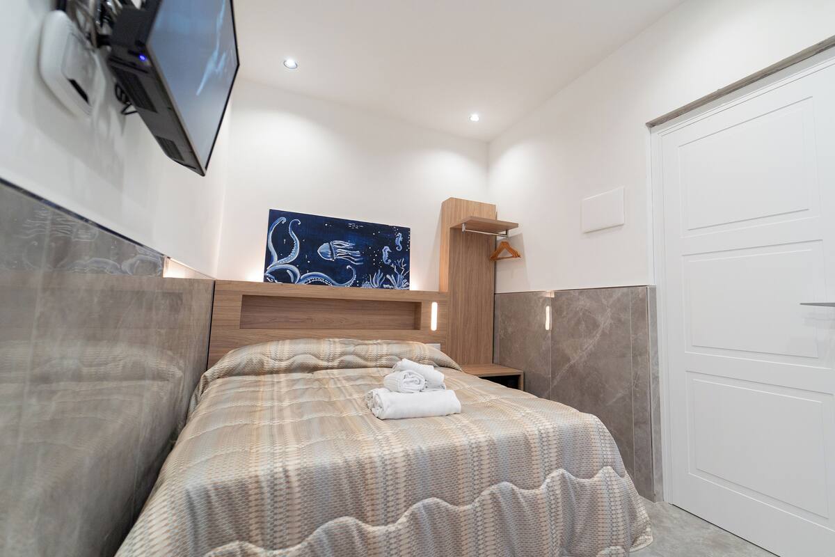 A bedroom with a bed, a TV mounted on the wall, a painting, and a door.
