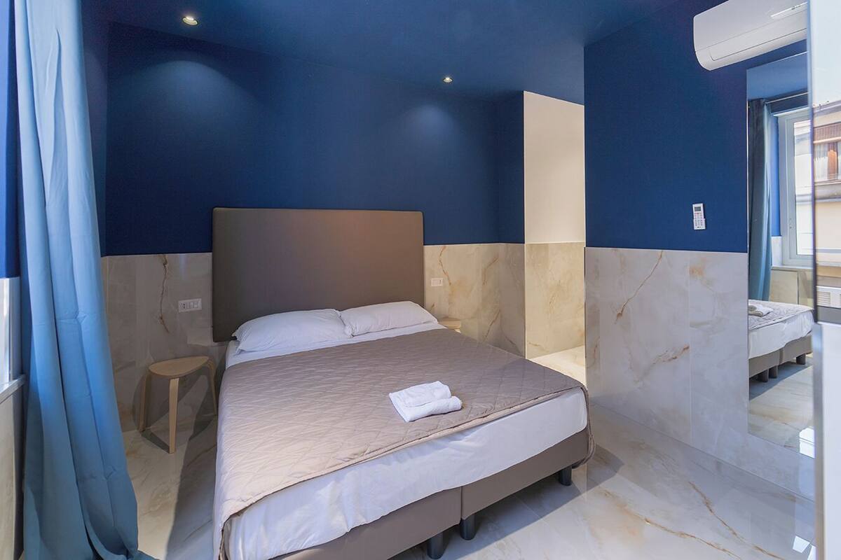 A hotel room with a bed, a small stool, a mirror, and a window.
