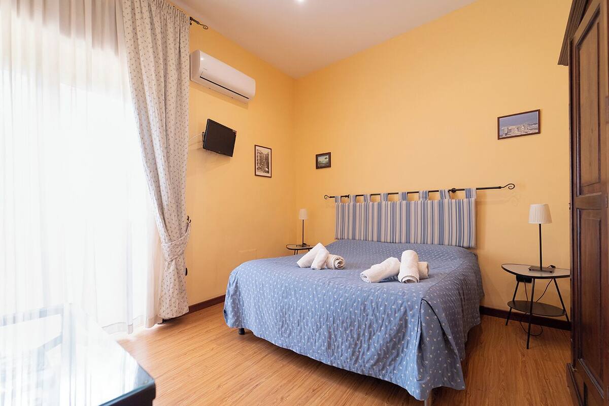 A hotel room with a bed, two bedside tables, a window with curtains, an air conditioning unit, and two framed pictures on the wall.
