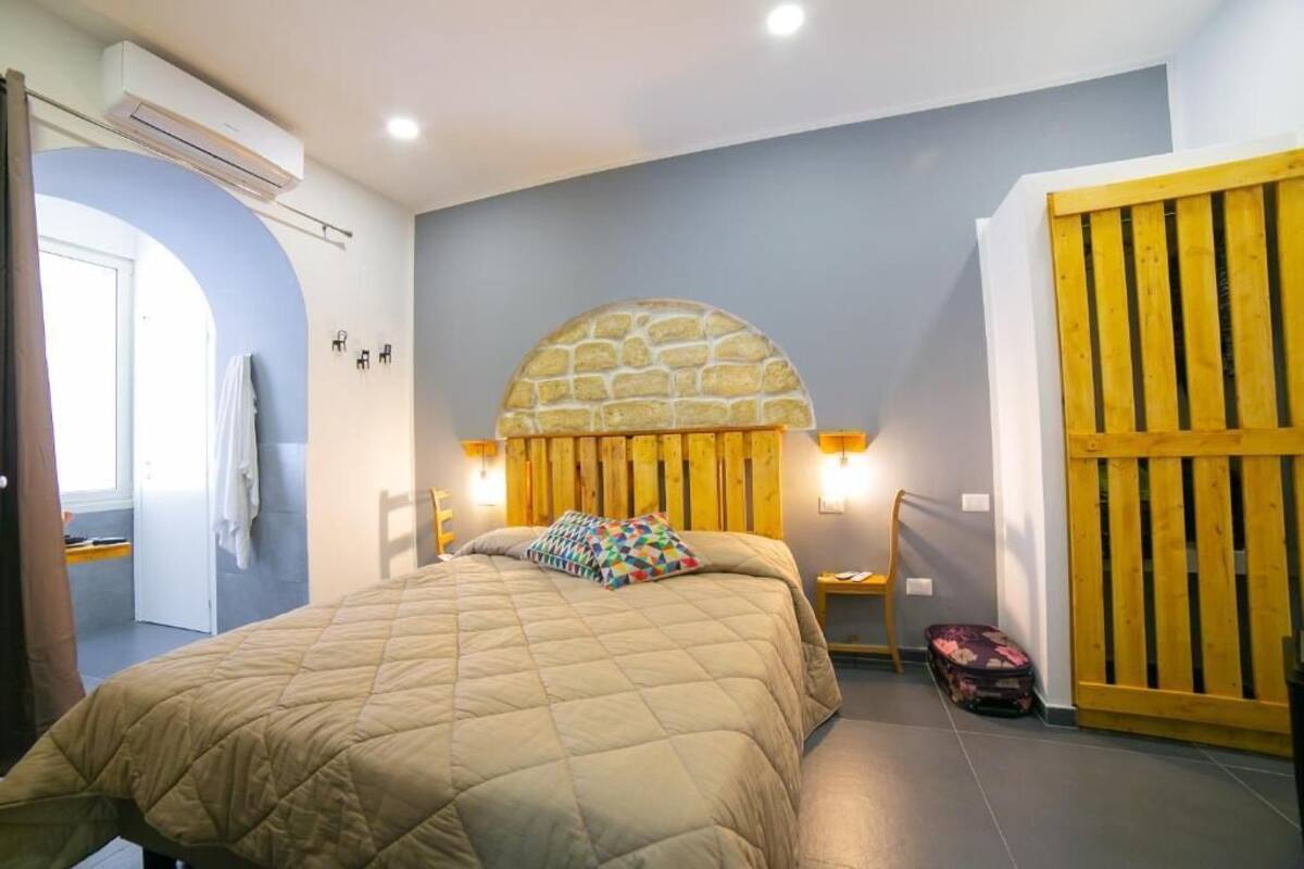 A bedroom with a stone arch headboard, a bed with a quilt, two bedside lamps, a wooden wardrobe, and a suitcase on the floor.