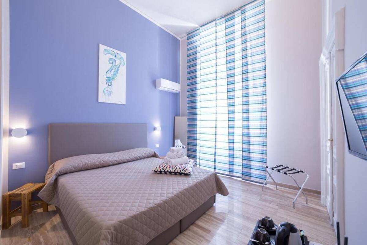 A bedroom with a bed, a bedside table, a wall-mounted air conditioner, a window with striped curtains, and a towel rack.