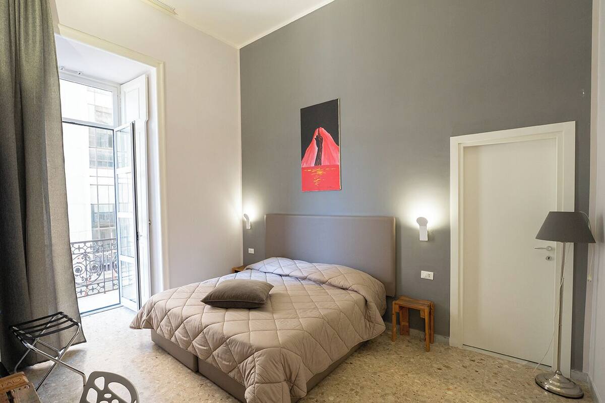 A bedroom with a bed, a small bedside table, a lamp, a door, and a window with a view of buildings.