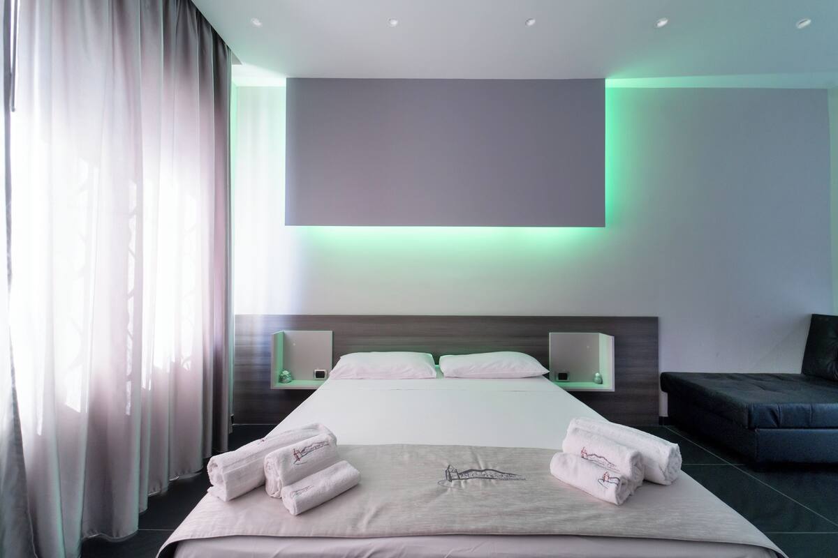 A modern hotel room with a large bed, a flat-screen TV mounted above it, and a bedside table with a lamp.