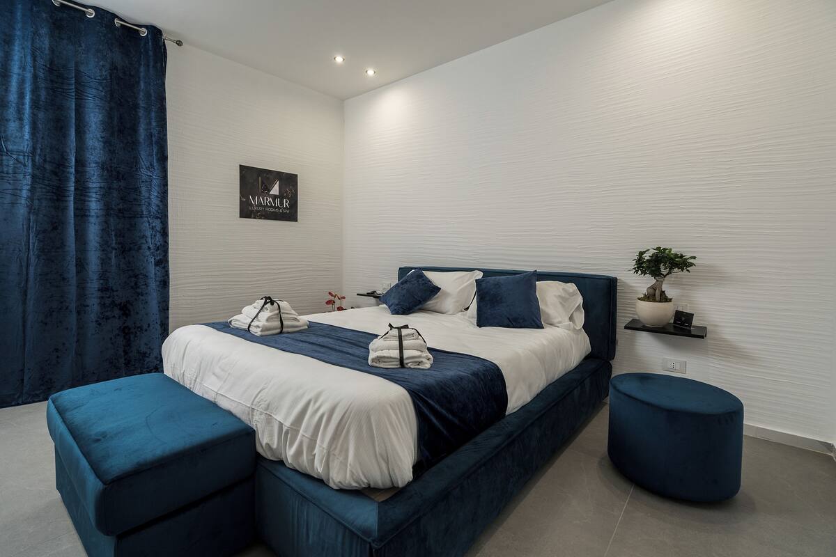A modern bedroom with a large bed, a bedside table, a small potted plant, and a blue curtain.