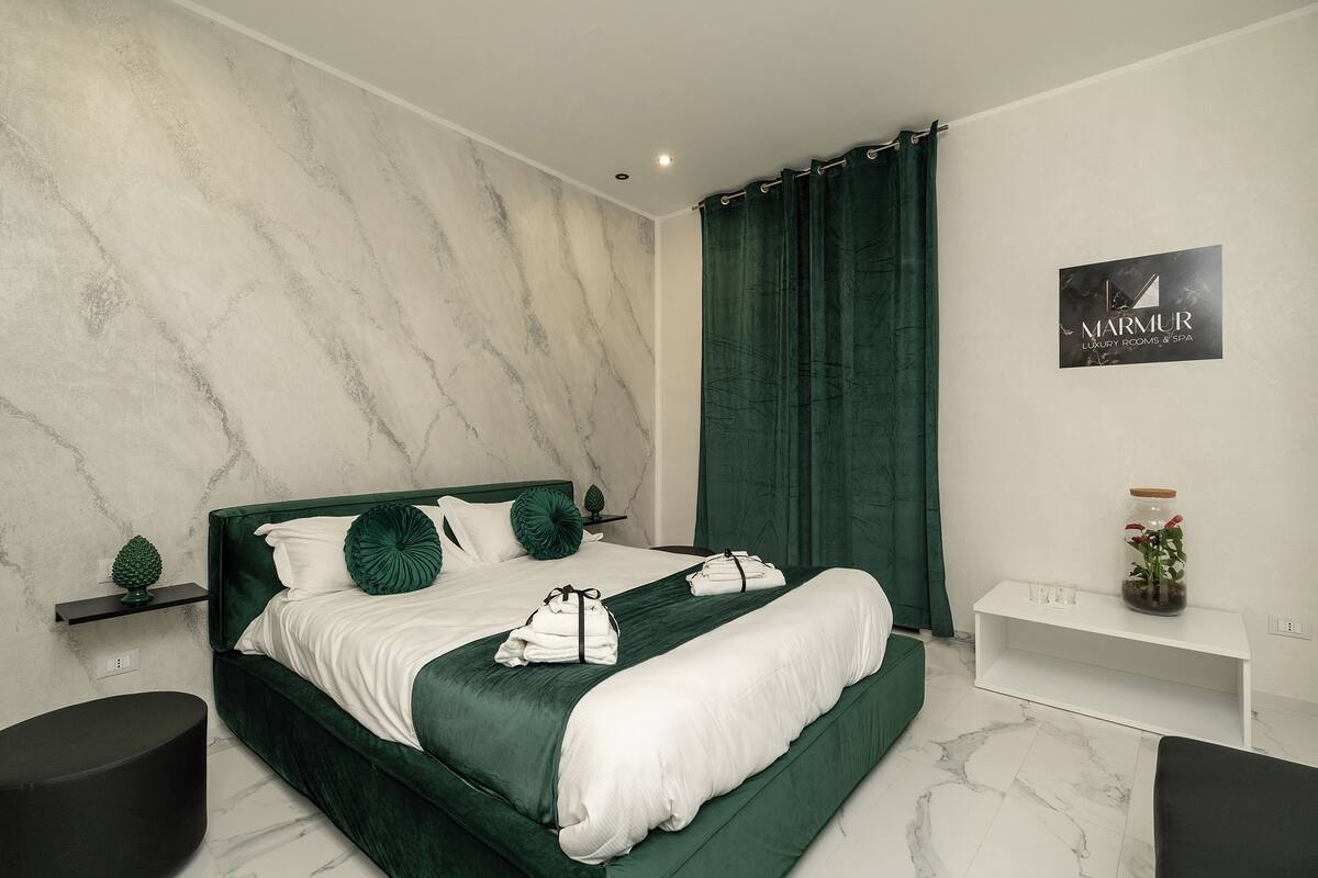 A modern bedroom with a large bed, green bedding, a marble wall, and a sign for 'Marmur Luxury Rooms & Spa'.