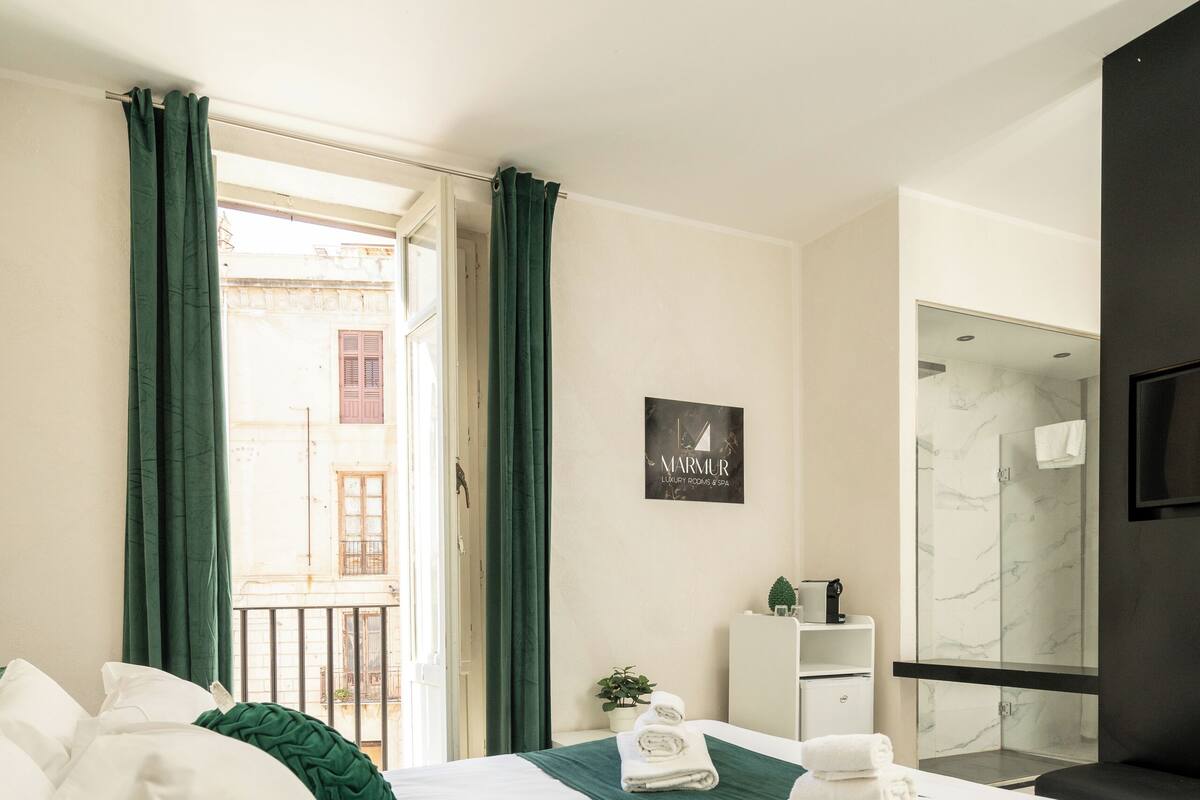 A modern hotel room with a large bed, a small desk, a shower area, and a view of the outside.