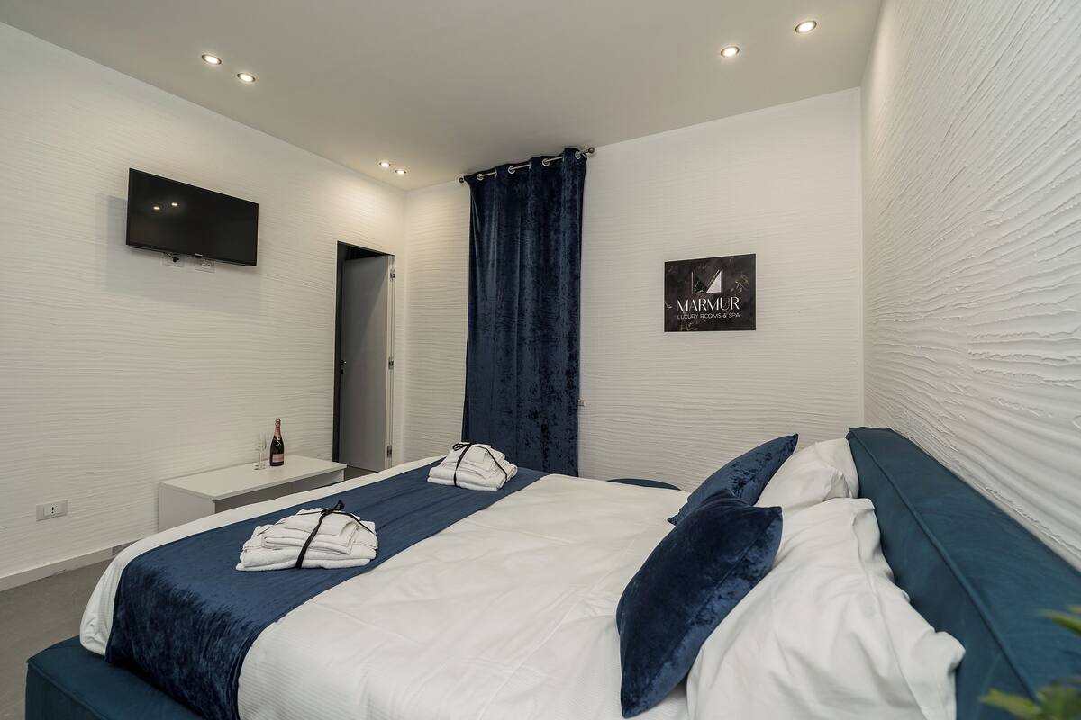 A modern hotel room with a large bed, a bedside table with a bottle and glasses, a flat-screen TV mounted on the wall, and a sign indicating the hotel name.